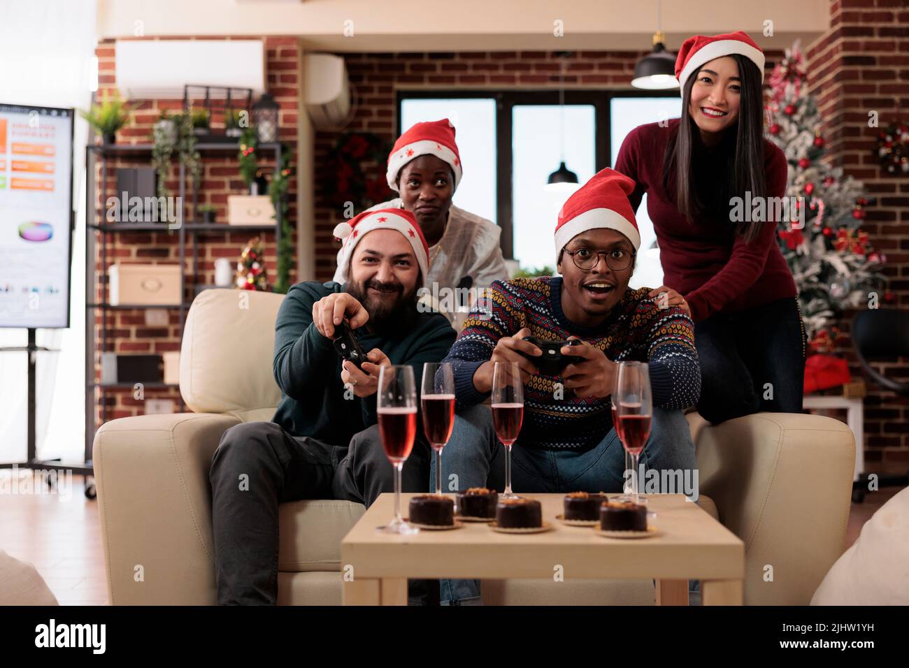 Multiethnic group of people playing video games, wearing santa hat at festive office party. Celebrating christmas eve festivity in workplace with xmas decorations, having fun with gaming console. Stock Photo