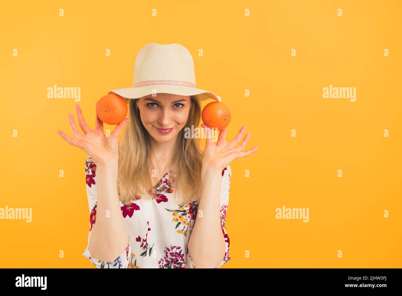 European thin blonde attractive woman in her mid 20s wearing a hat and a floral blouse showing to the camera two juicy oranges over orange background. High quality photo Stock Photo