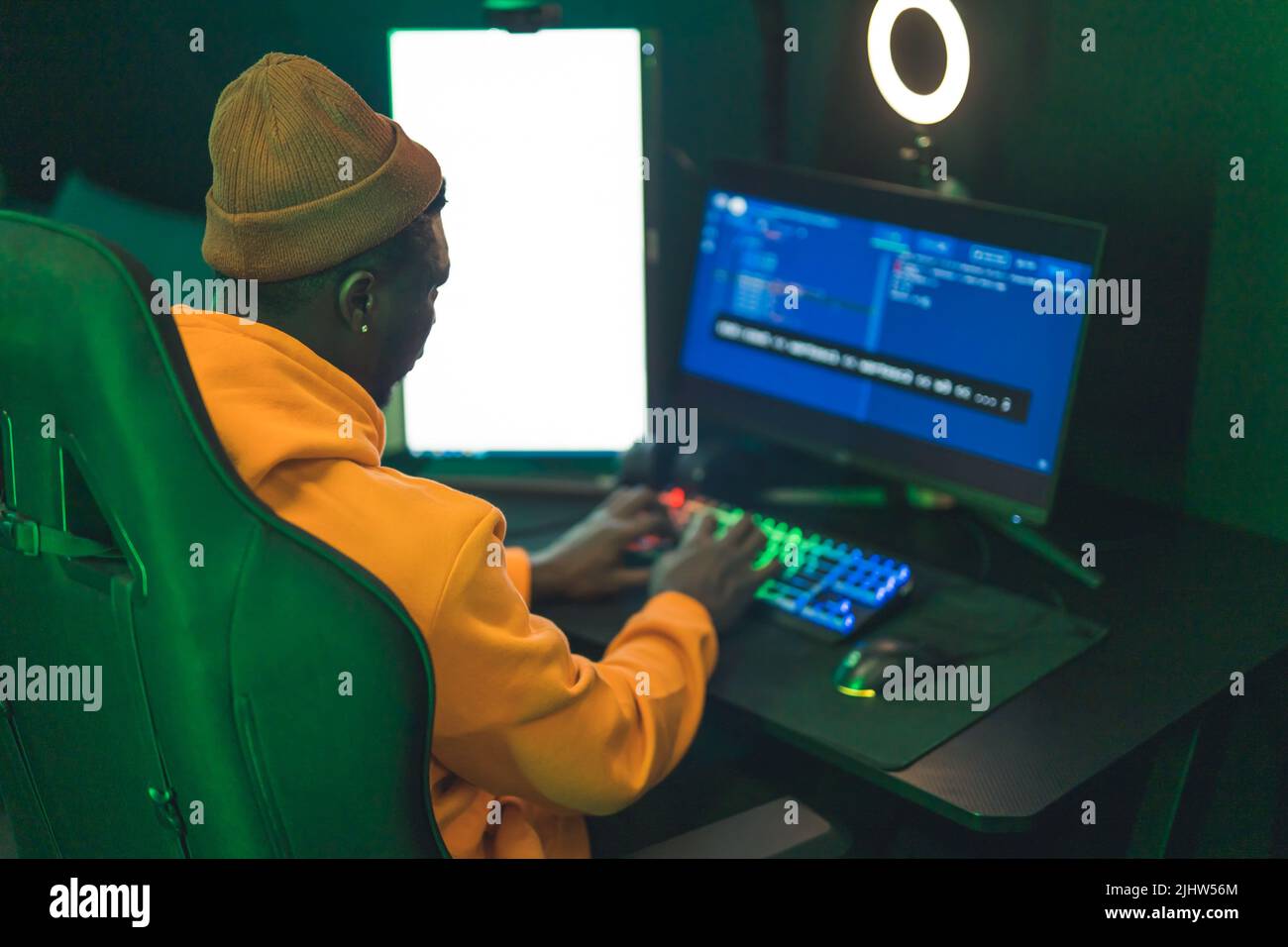 Back view of an african american wearing an orange hoodie and hat sitting on a tall chair, watching a computer screen. . High quality photo Stock Photo