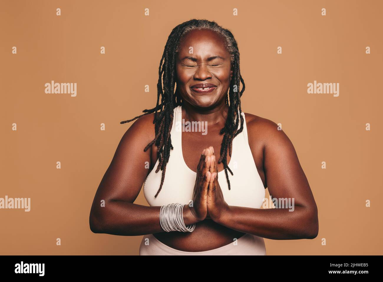 Cheerful woman meditating with her eyes closed and her hands in prayer position. Happy woman with dreadlocks practicing yoga in a studio. Sporty matur Stock Photo