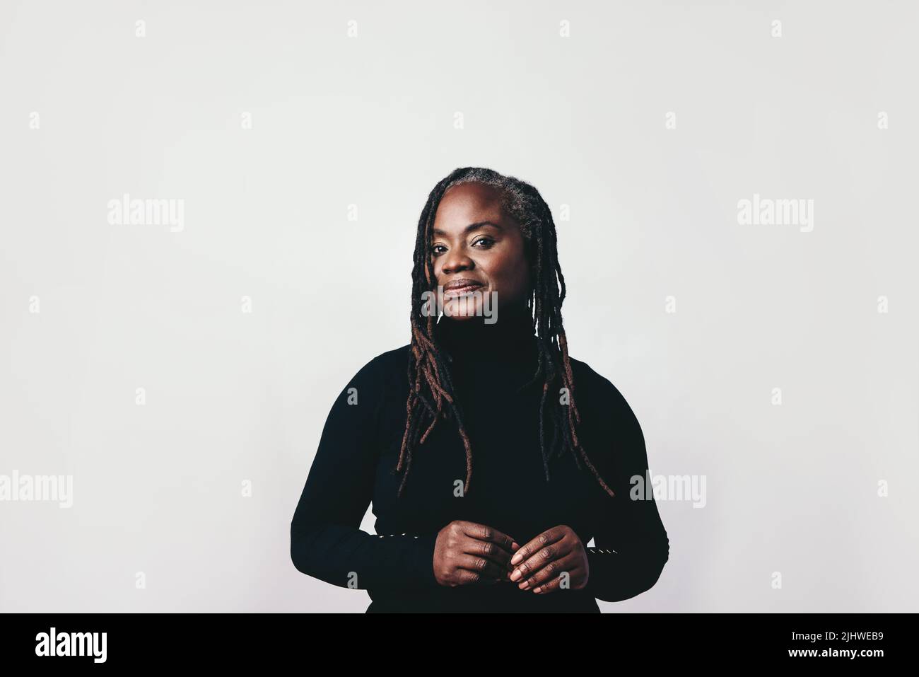 Confident middle-aged woman with dreadlocks looking at the camera in a studio. Elegant black woman standing against a grey background. Stock Photo