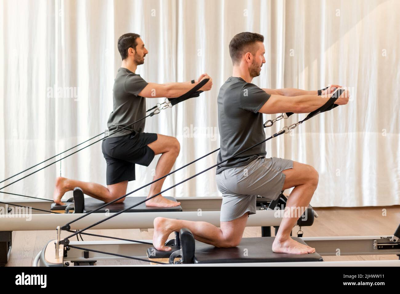 Full body side view of strong men in sportswear doing hug tree kneeling pilates exercise with resistance bands on reformer bed during training in gym Stock Photo