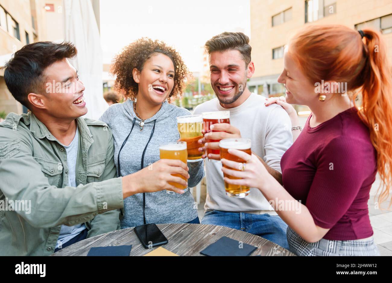 Positive multiethnic friends clinking glasses of beer and looking at each other while sitting together at table in outdoor pub on summer day in city Stock Photo