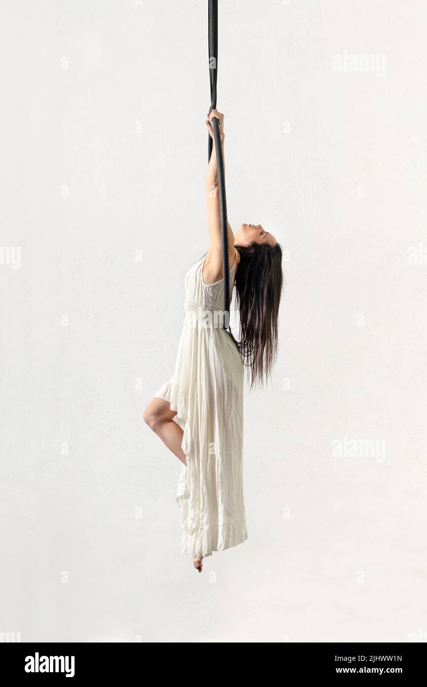 Side view of barefoot brunette in long dress hanging on aerial silks while doing gymnastic against white background Stock Photo