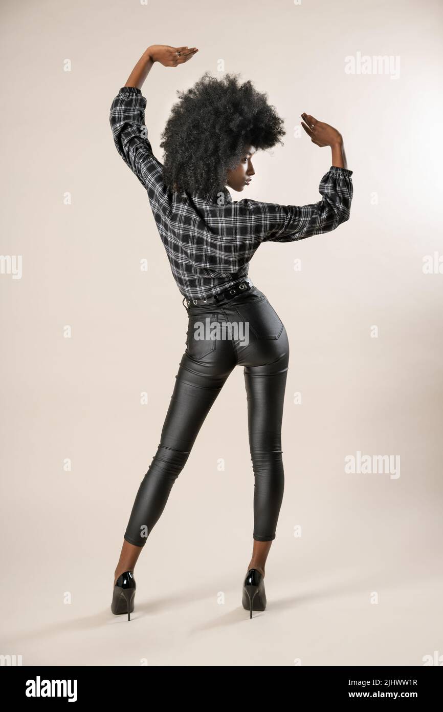 Back view of curly black woman in checkered shirt and black trousers standing on high heels against beige background Stock Photo