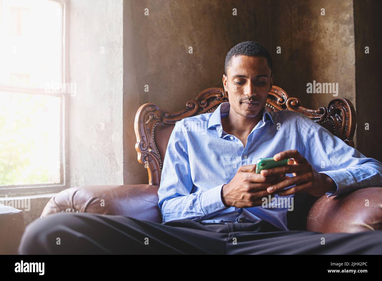 A young African American man in a shirt types on his smartphone sitting in a comfortable armchair in the living room Stock Photo