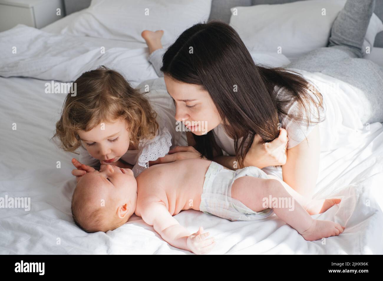 Older sister, mother and baby brother communicating. Two cute little Caucasian children siblings playing at home. Adorable smiling family lying Stock Photo