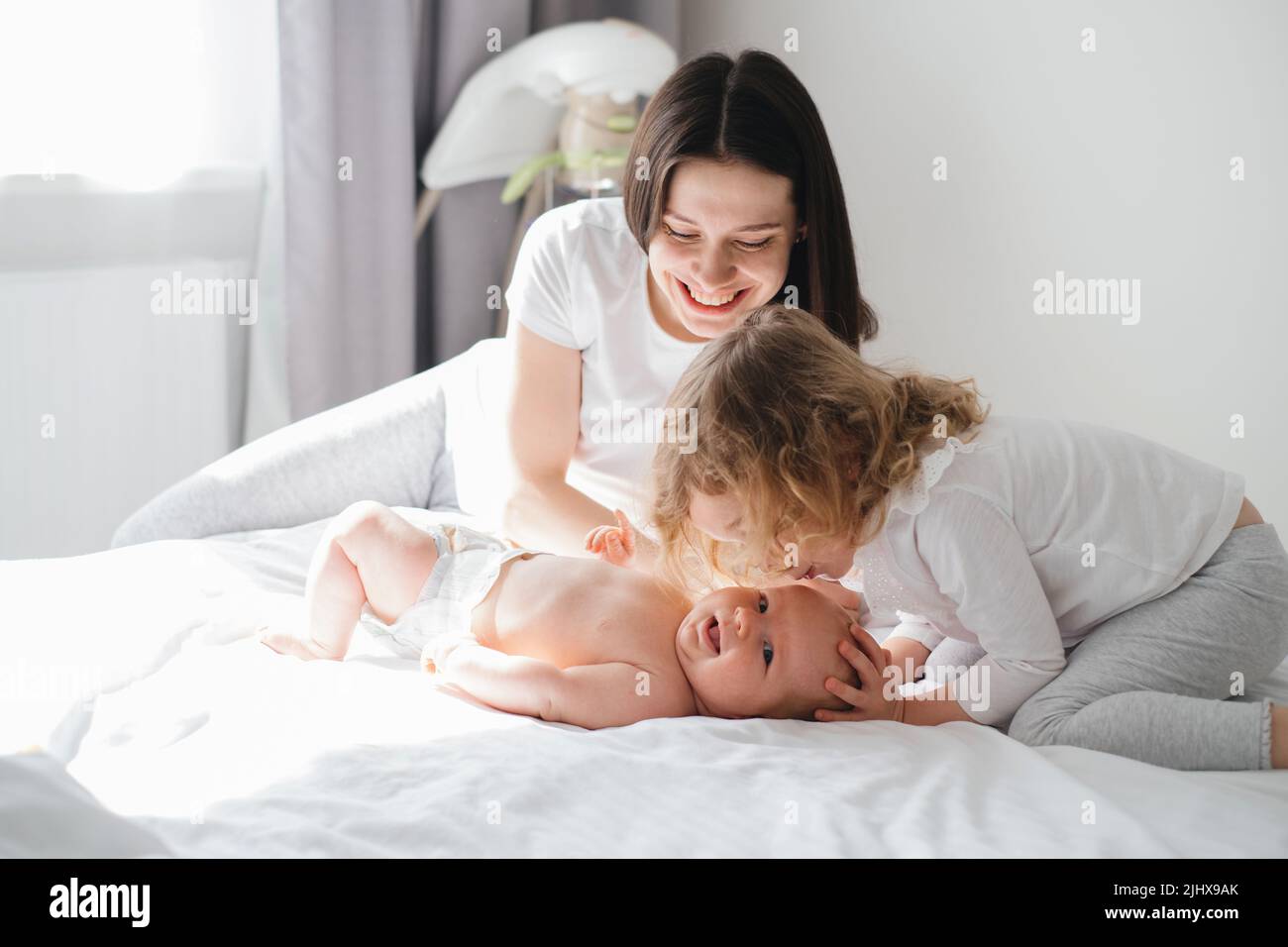 Older sister, mother and baby brother communicating. Two cute little Caucasian children siblings playing at home. Adorable smiling family lying Stock Photo