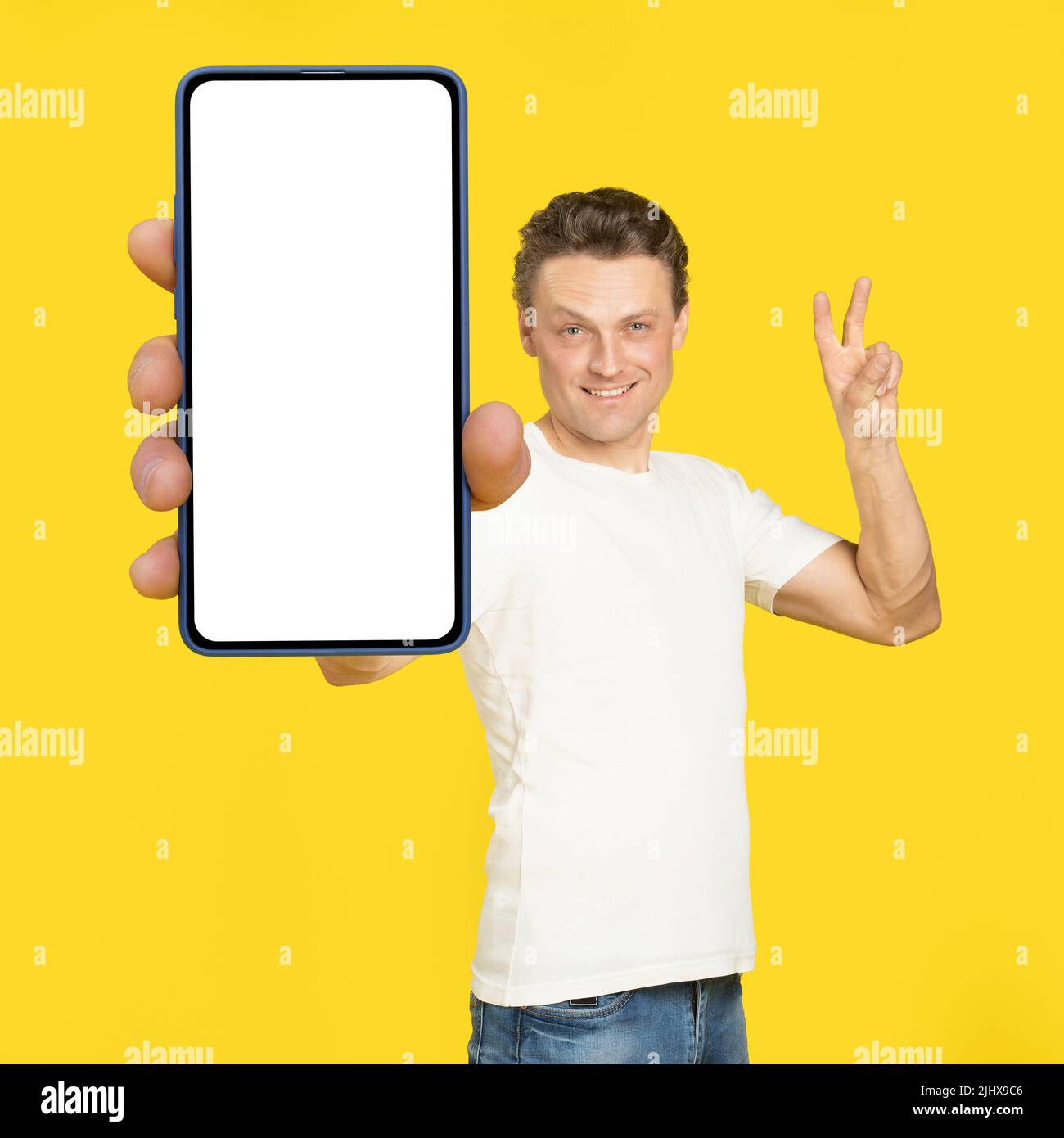 Gesturing V or victory handsome man holding huge smartphone with white screen wearing white t-shirt and jeans isolated on yellow background. Mobile app advertisement, great offer. Stock Photo