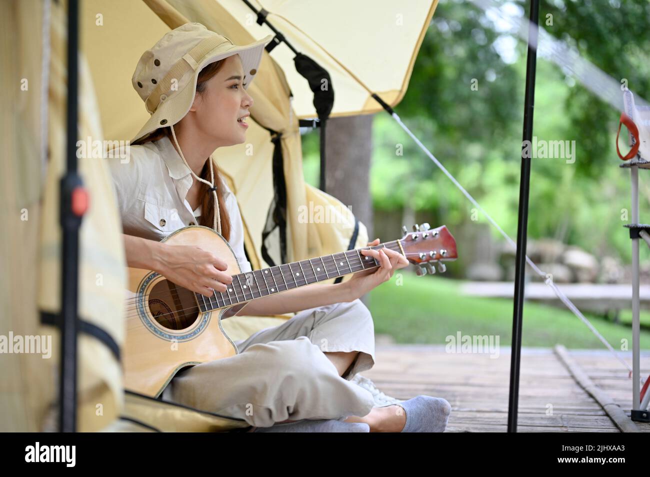 Beautiful charming young Asian woman sing a song while playing guitar in her minimal camping tent. Camping and outdoor activity concept. Stock Photo