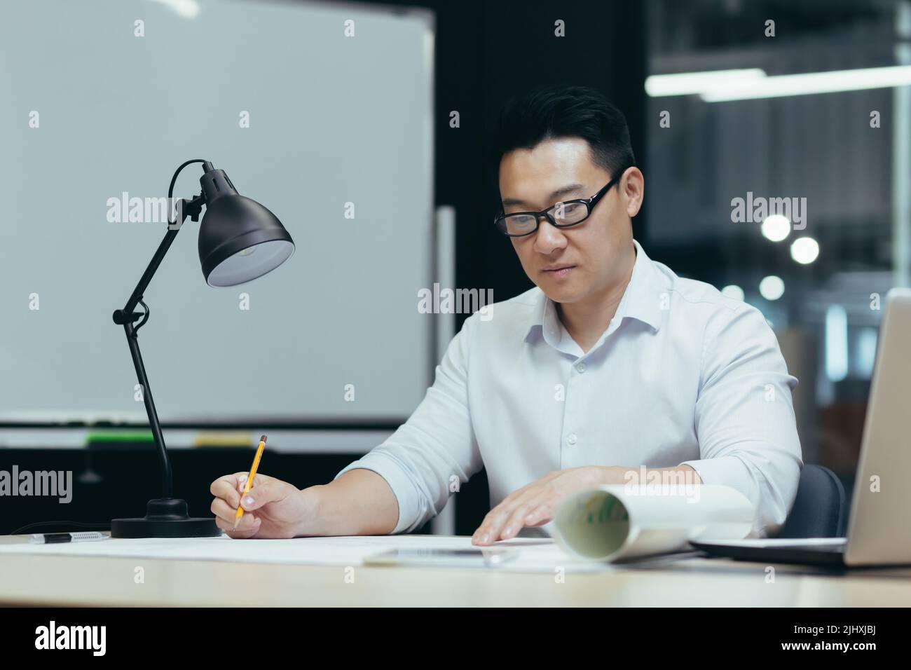 Asian designer working on blueprint in modern office, male architect thoughtfully working on construction project Stock Photo