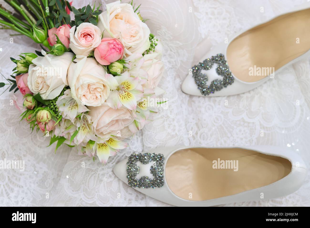 Wedding day composition with silk white shoes, pastel bridal bouquet on white lace blurry background. Top view Stock Photo