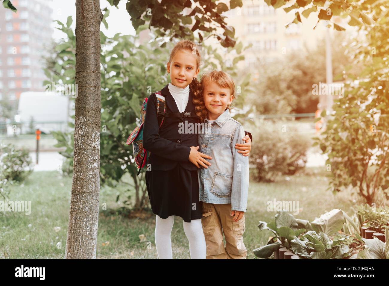 back to school. little happy kid pupil schoolgirl eight years old in fashion uniform with backpack and her preschool brother boy hug together ready go Stock Photo