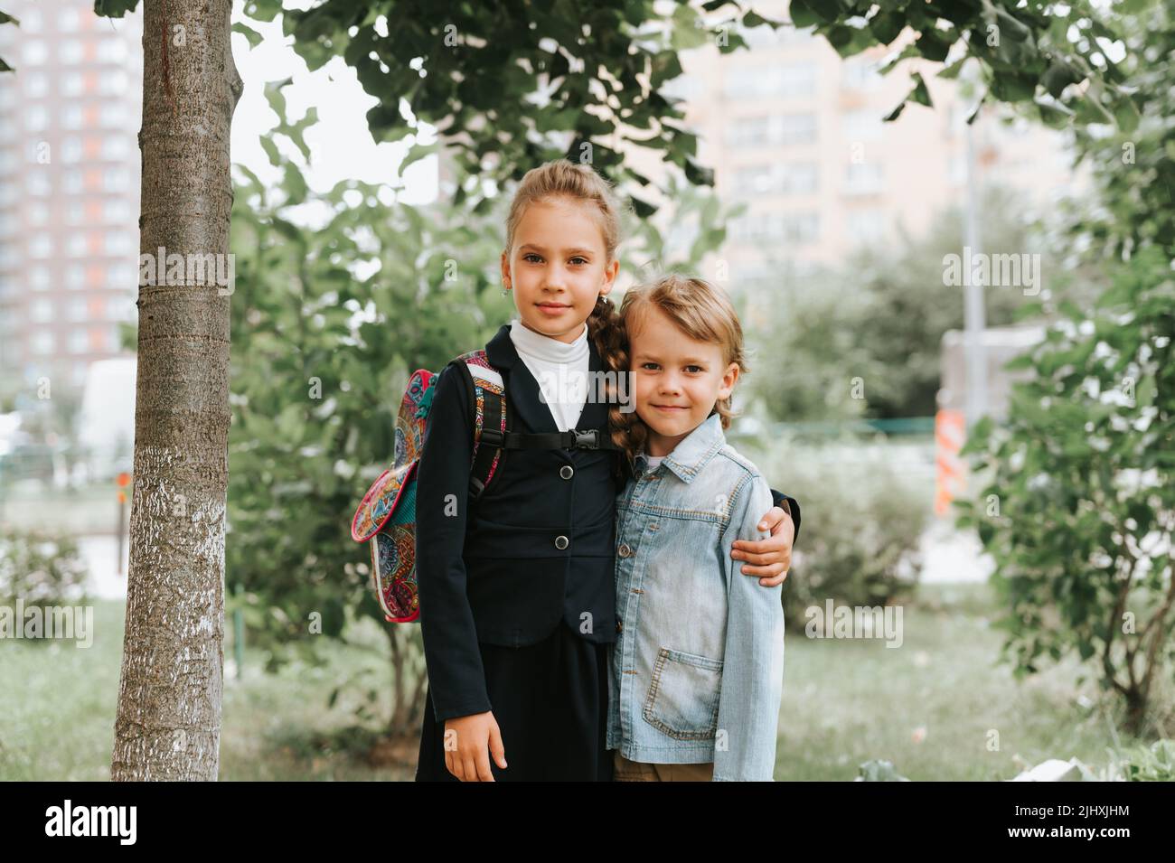 back to school. little happy kid pupil schoolgirl eight years old in fashion uniform with backpack and her preschool brother boy hug together ready go Stock Photo