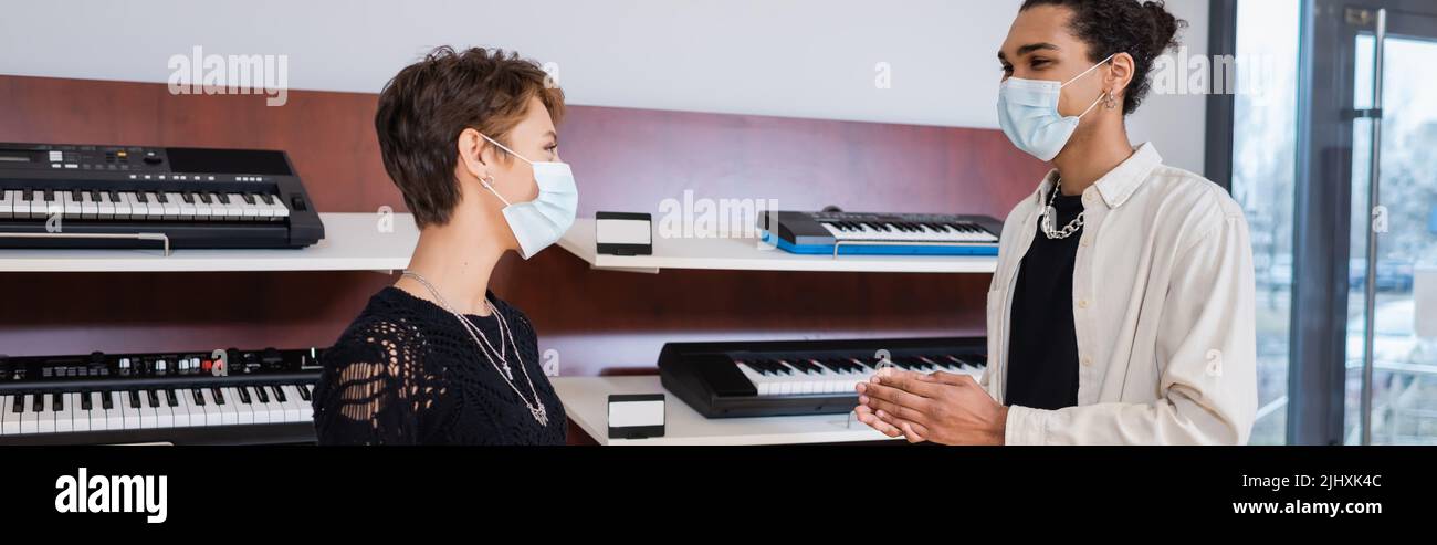 African american seller and customer in medical masks standing near synthesizers in music shop, banner Stock Photo