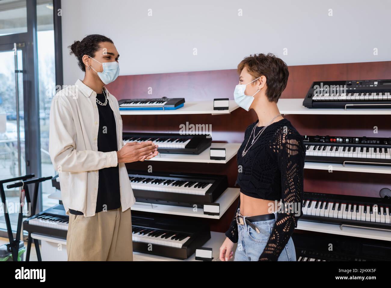 African american seller in medical mask looking at customer near synthesizers in music store Stock Photo
