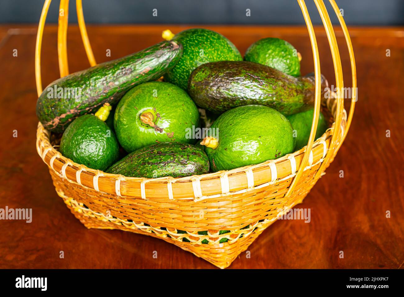 Pile of different varieties of fresh green avocado in wicker bamboo basket on wooden table. Stock Photo