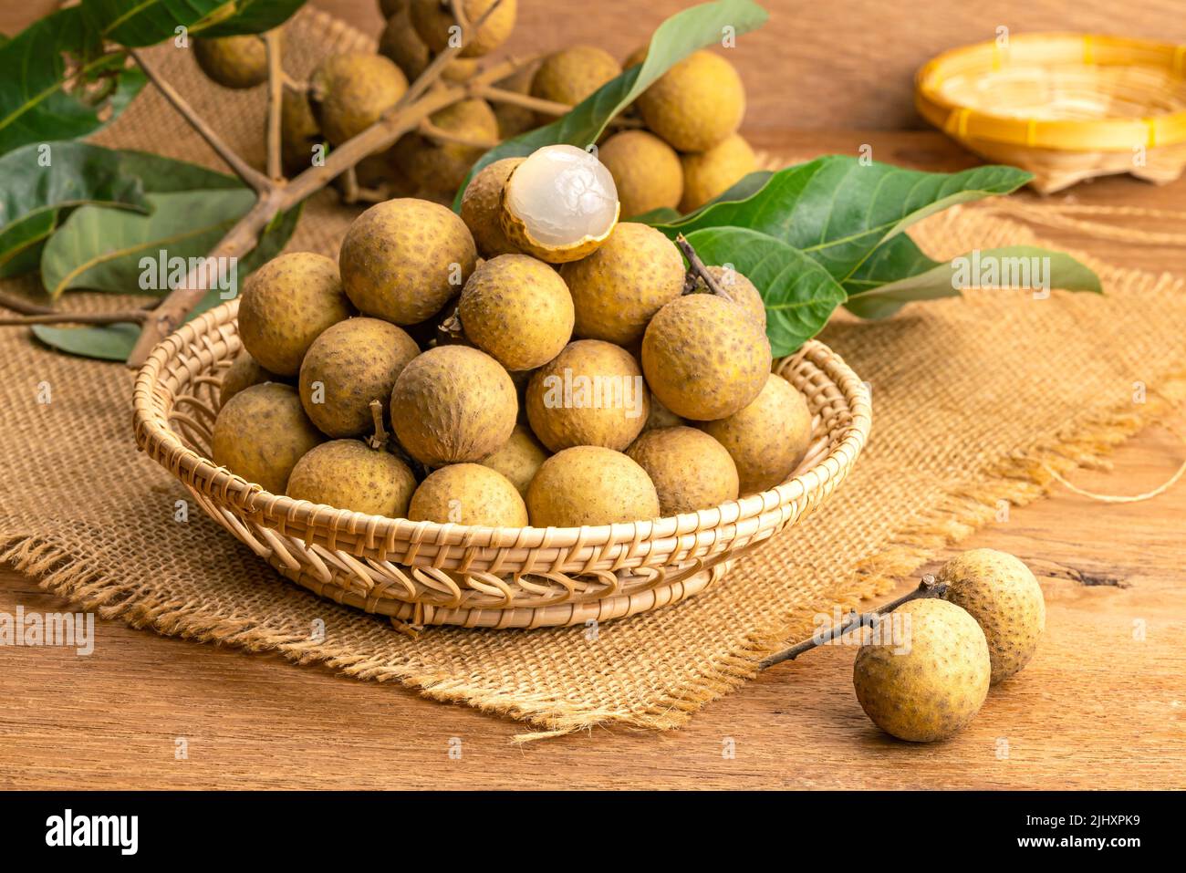 Pile of fresh langan in bamboo basket with bunch of longan and green leaves place on sack cloth on wooden table. Stock Photo