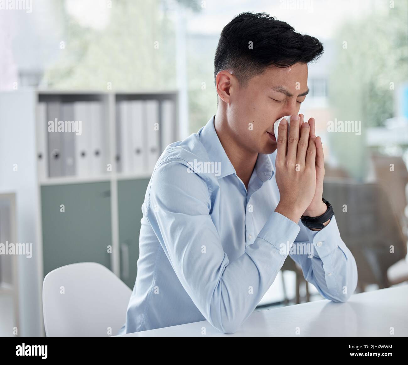 A sick businessman using a tissue and blowing his nose while working in a modern office. An ill asian man ill with flu, a cold or covid. Male employee Stock Photo