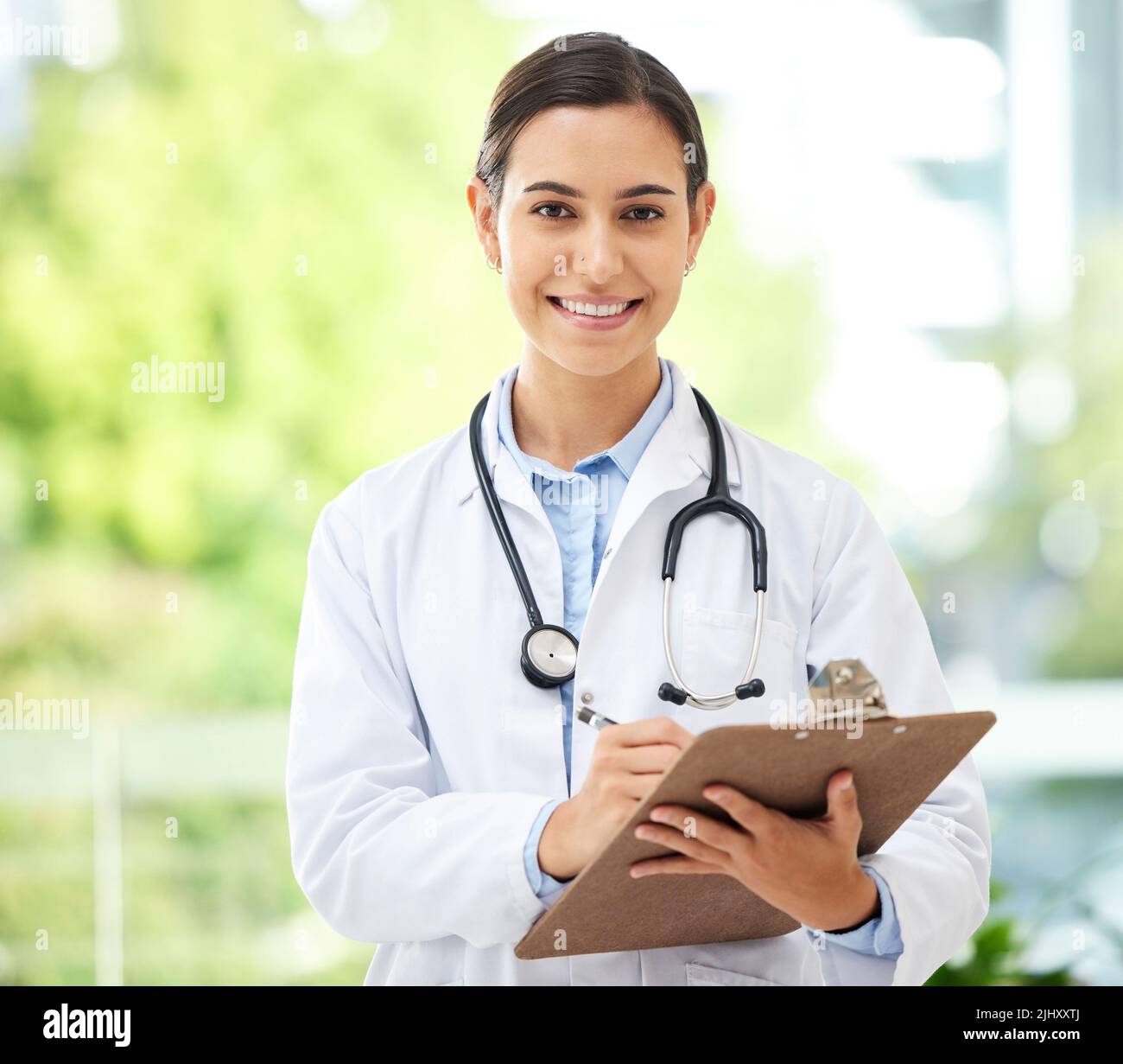 Confident young mixed race female doctor standing with clipboard a medical office. One hispanic woman in a white coat with stethoscope. Trusted Stock Photo