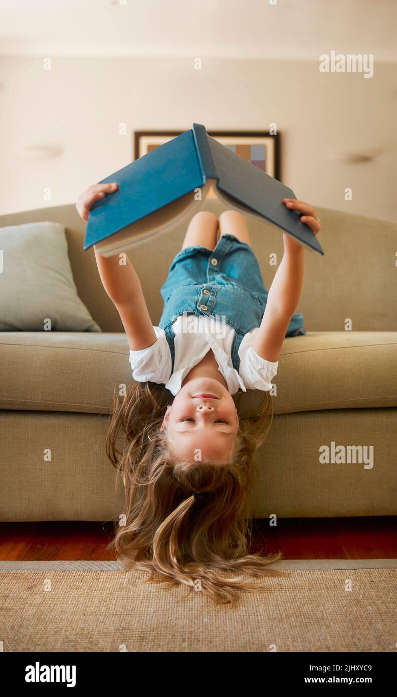 Carefree content caucasian little girl reading a book alone sitting upside down on the couch at home. One cheerful positive child holding and reading Stock Photo