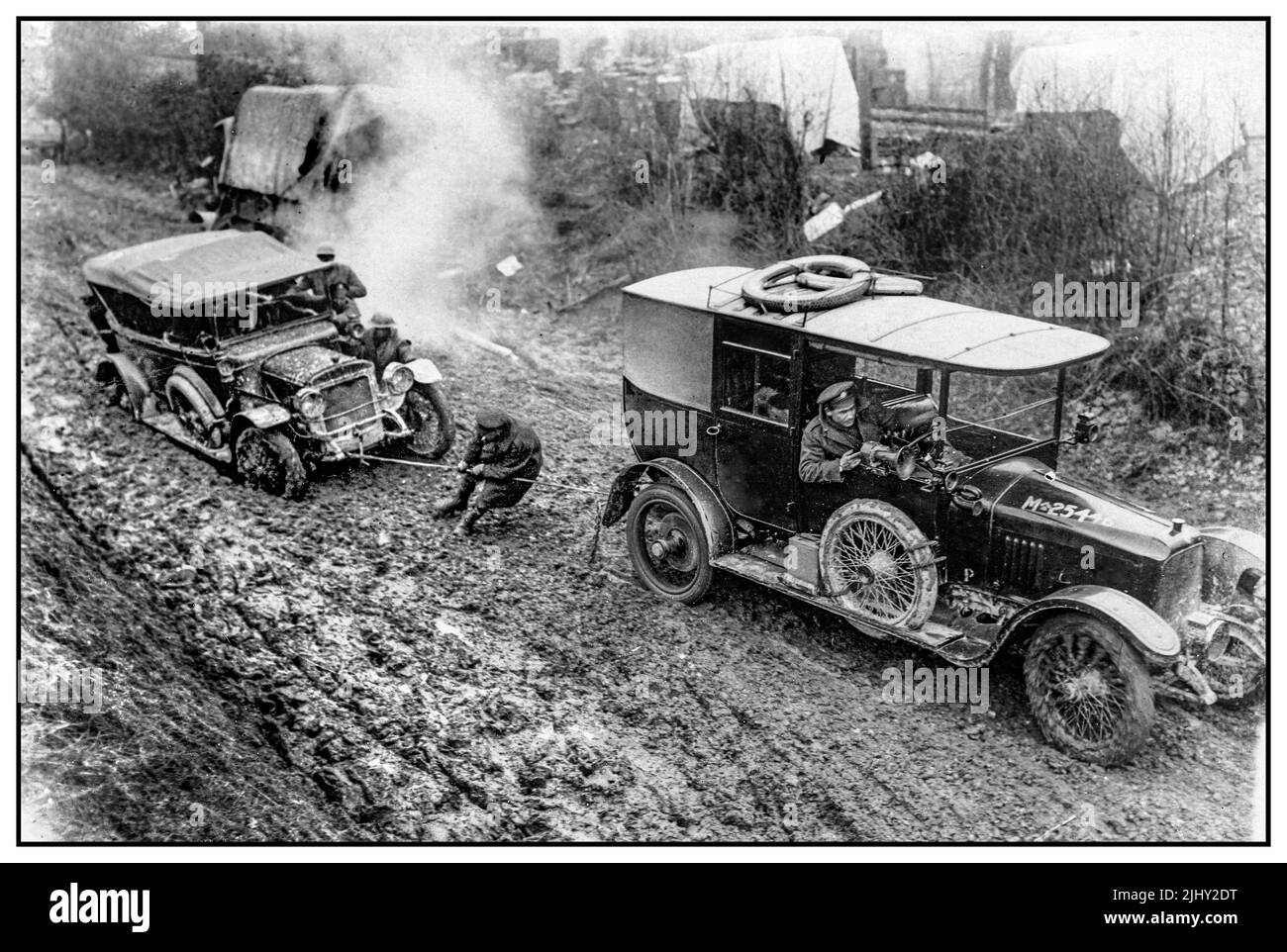 WW1 British Army on The Western Front in France with car being towed during World War I. This image shows a car being towed through thick mud. As the car in front tries to pull the other free, a soldier lends a hand by pulling on the tow rope. A group of soldiers standing next to the immovable car add their weight to the struggle. There appears to be a camp directly behind the road they are on. The car in front has the number 'M25446' painted on the bonnet. Extreme weather conditions, floods and deep mud that ensued, created tremendous logistical problems and hazards for the soldiers at the We Stock Photo