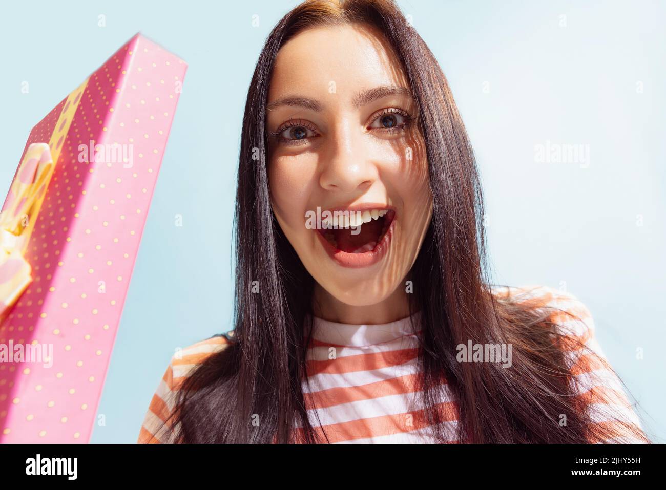 Closeup face of young beautiful astonished girl holding gift box isolated on blue background. Concept of emotions, holidays, inspiration, sales, ad. Stock Photo