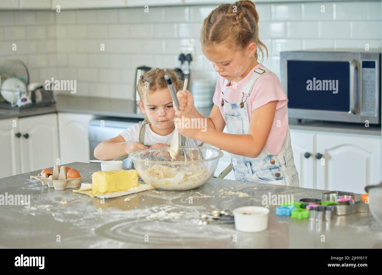 Two messy little girls baking in the kitchen at home. Caucasian focused sisters learning to make a dessert and cook. Adorable children bonding and Stock Photo