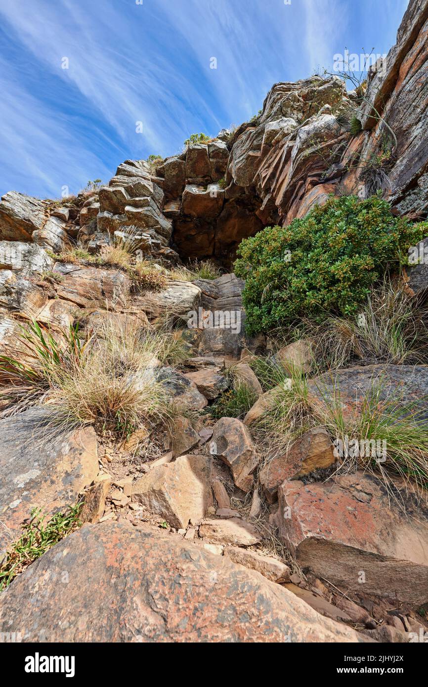 A rocky mountain with plants and shrubs growing against a cloudy sky background with copy space. Rugged, remote and quiet landscape with rocks and Stock Photo
