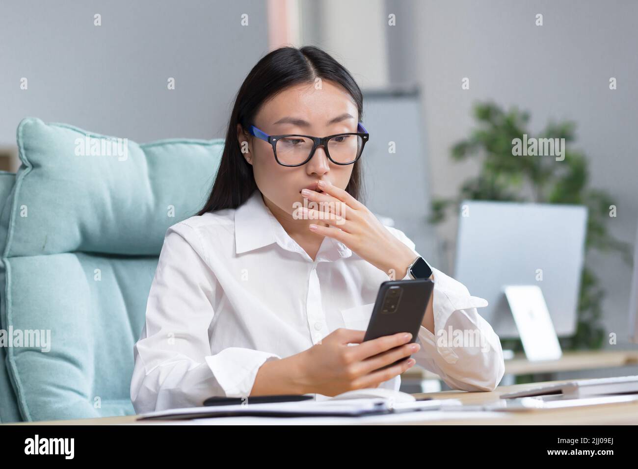 Worried young beautiful Asian business woman in glasses holds the phone in her hands, looks at the phone, received bad news, surprised, shocked. Sitting at a desk in a modern office. Stock Photo