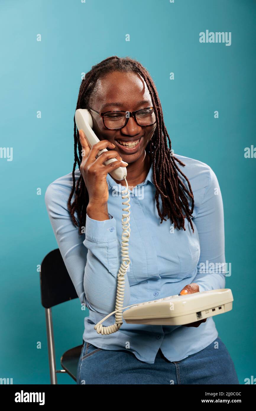 Cheerful smiling heartily young woman answering friend calling on landline phone device on blue background. Positive african american helpline operator talking on wired telephone. Stock Photo