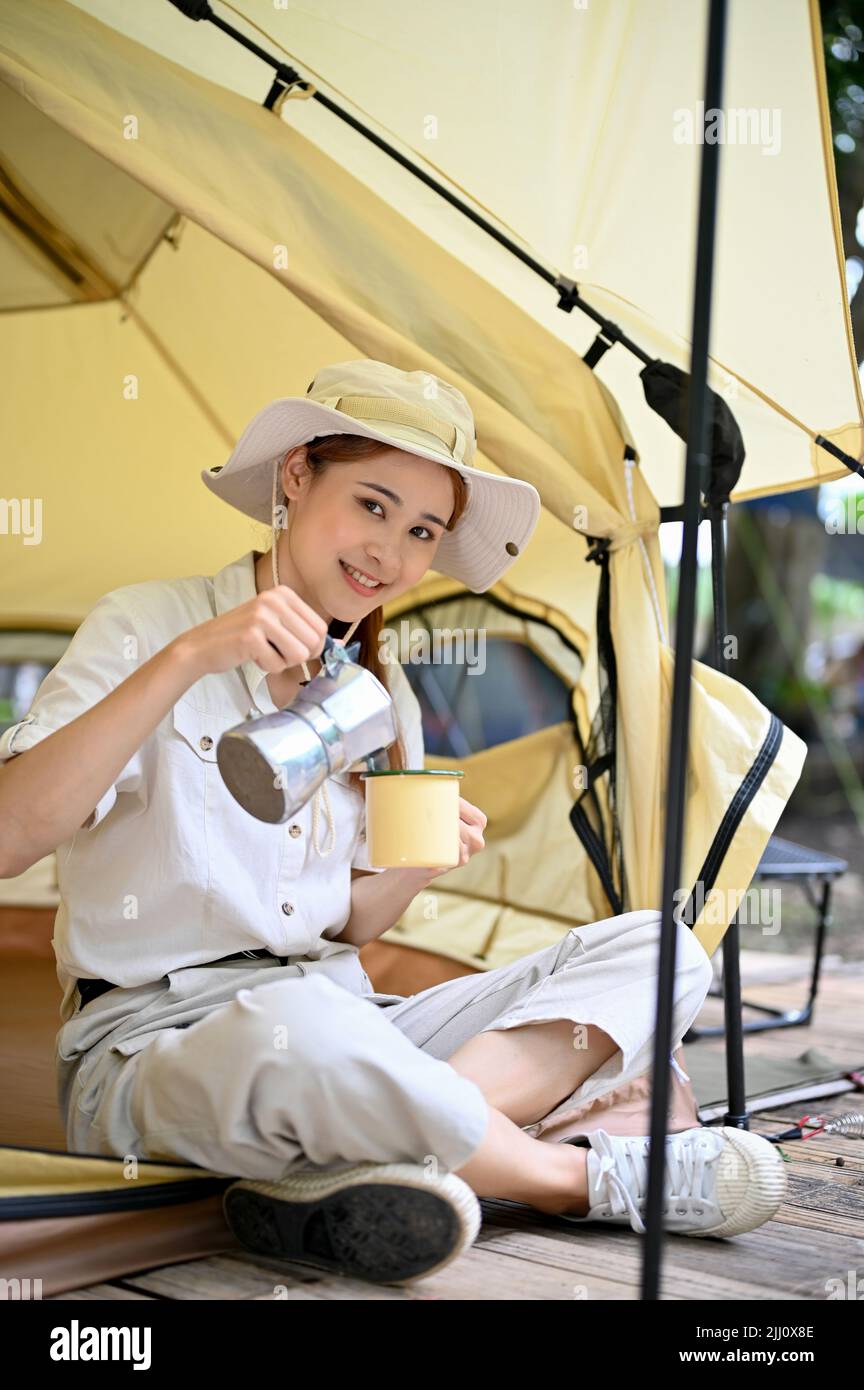 Attractive charming young Asian female camper pouring a hot coffee into a coffee cup, having a coffee in the morning in front of her tent. Stock Photo