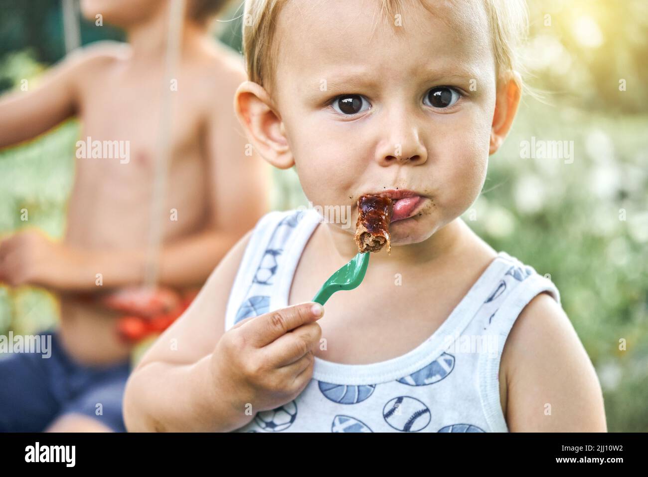 Cute blond toddler boy eats barbecue sausage with plastic fork on blurred background. Portrait of little boy with dark brown eyes at picnic on nature Stock Photo