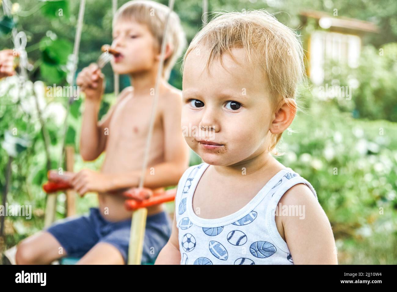 Brothers enjoy barbecue sausage at picnic on nature with lush greenery in countryside. Portrait of cute blond toddler boy on blurred background Stock Photo