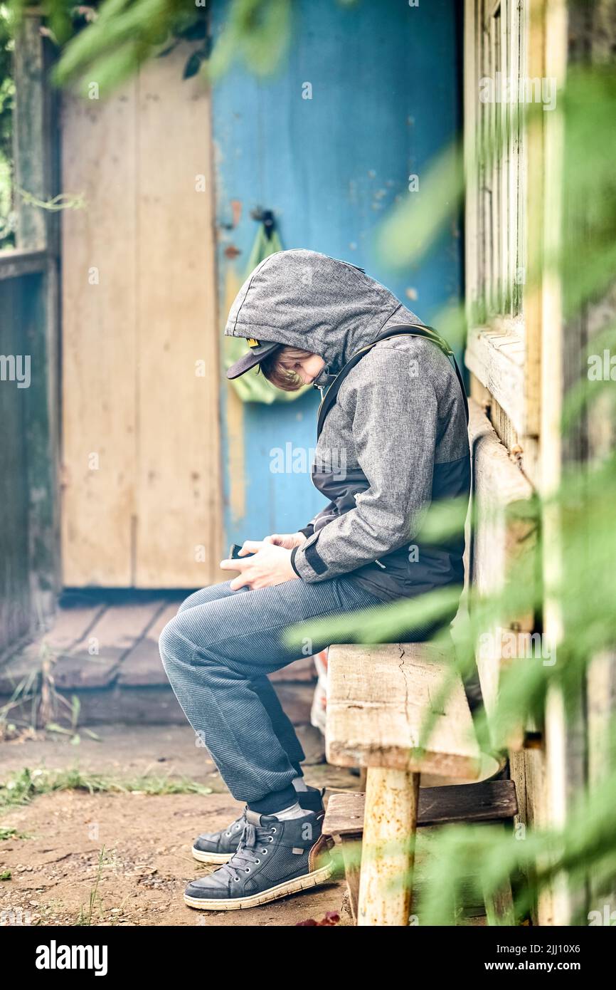 Focused schoolboy in warm jacket uses smartphone sitting on wooden bench near house in countryside. Boy plays game on phone after school in yard Stock Photo