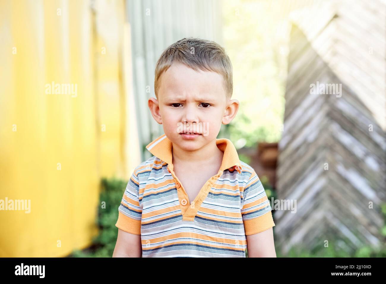 Cute toddler with blond hair frowns standing in yard on blurred background. Little boy spends summer vacation in countryside on sunny day closeup Stock Photo