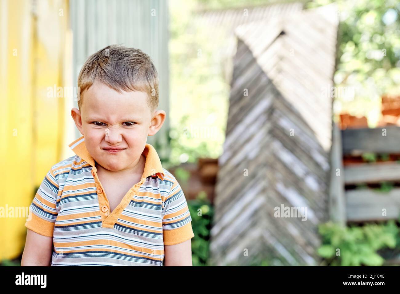 Little boy shows grimaces standing in yard on blurred background. Portrait of blond toddler against lush green bushes on sunny day in countryside Stock Photo