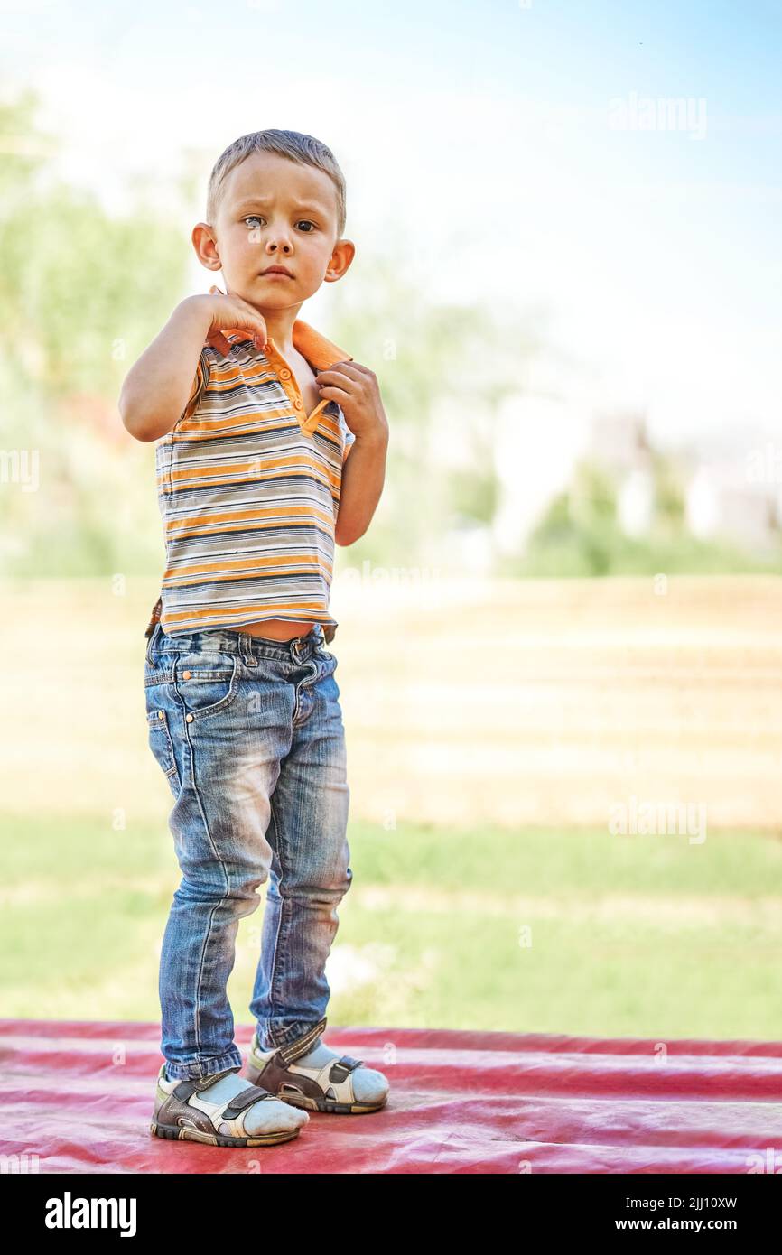 Serious little boy in jeans stands on red bench on blurred background. Cute toddler with blond hair enjoys sunny day in countryside Stock Photo