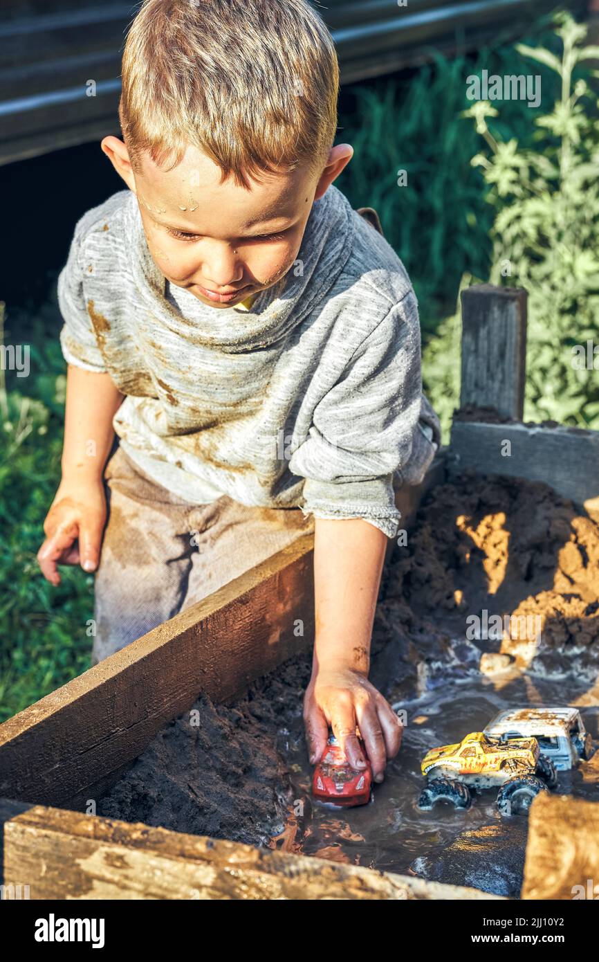 Cute serious preschooler boy with dirty face and clothes playing with toy cars in the sandbox near wooden rural house in summer day in countryside Stock Photo