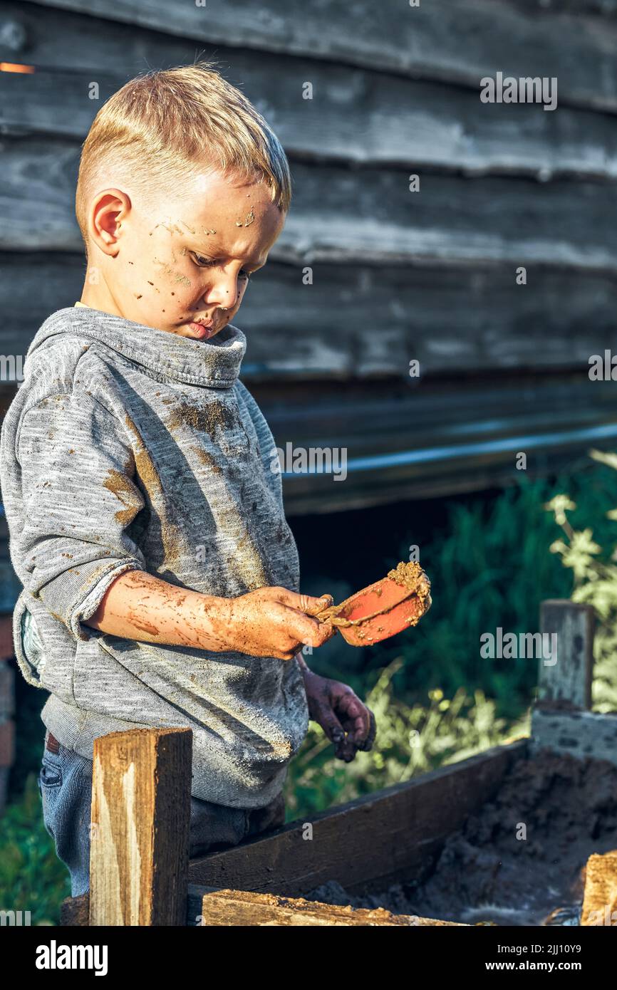 Cute serious preschooler boy with dirty face and clothes playing with toy cars in the sandbox near wooden rural house in summer day in countryside Stock Photo