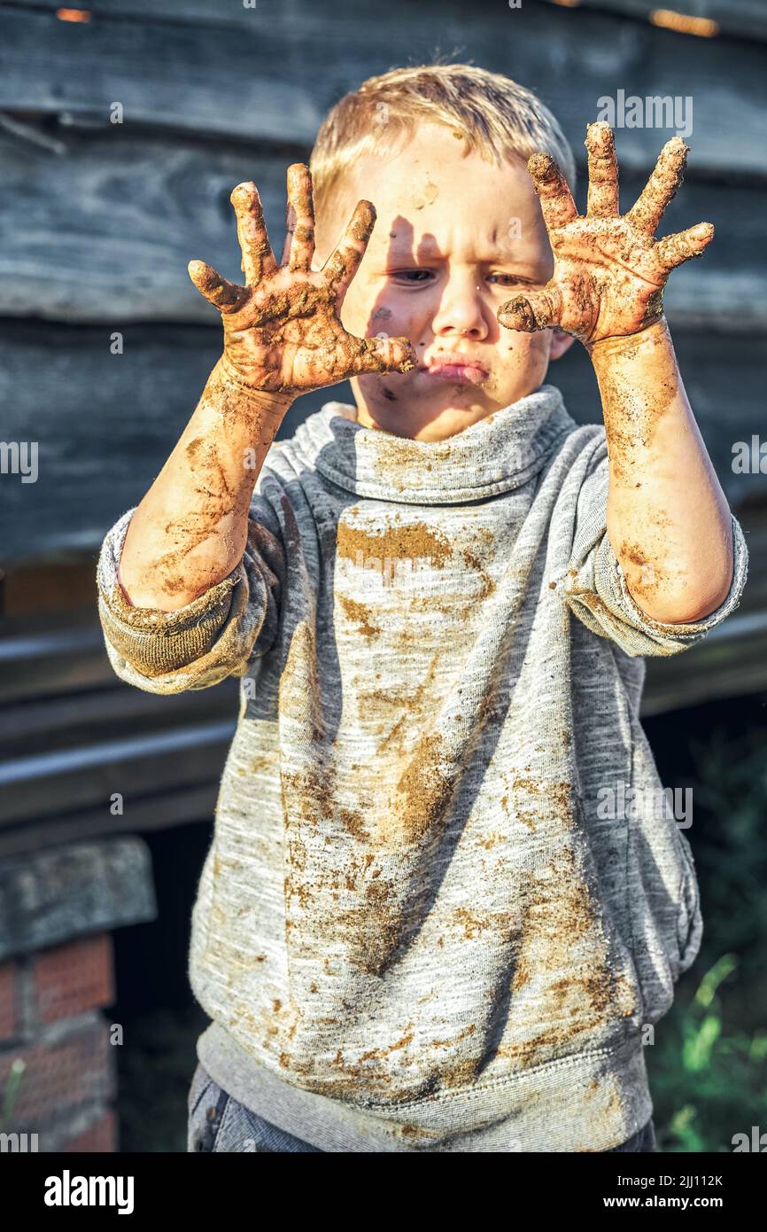Angry boy gets dirty after playing in the sandbox near wooden rural house in summer day in countryside Stock Photo