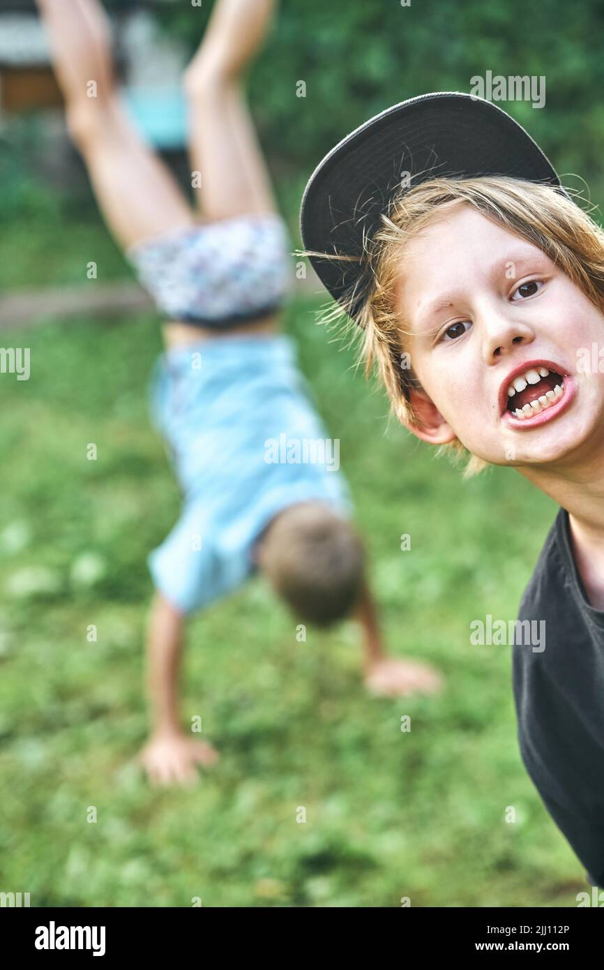 Cute boy in hat looks in camera against blurred view of elder brother making handstand on grass. Schoolboys have fun in countryside closeup Stock Photo