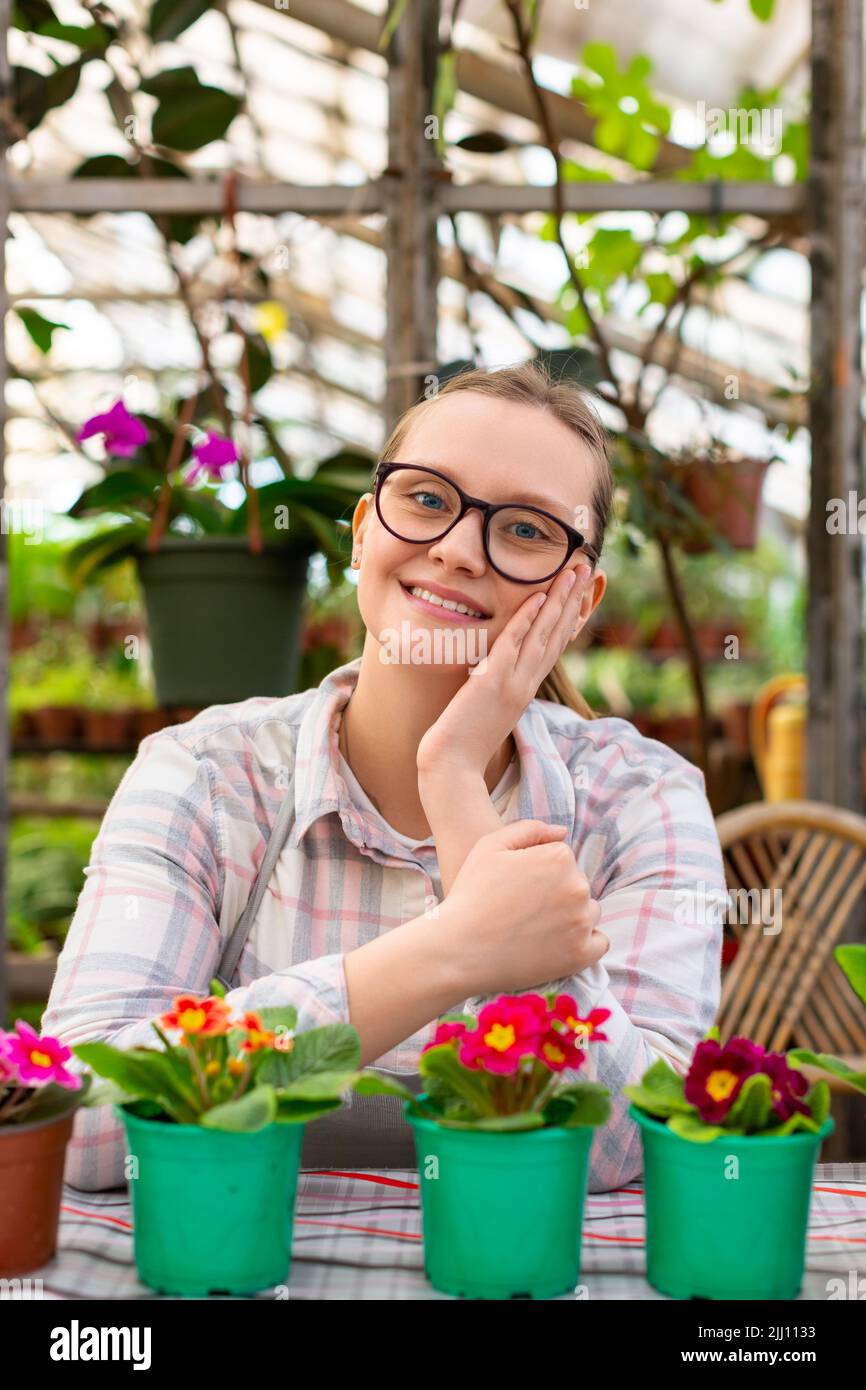A young woman with several flowers in green pots in a greenhouse looking at camera and smiling. Vertical photo. Stock Photo