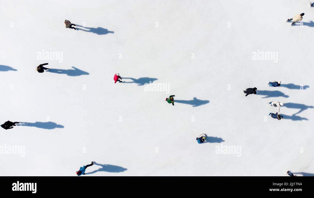 Many people are skating on a white outdoor ice rink in the city on a sunny winter day. Shadows of people skating on the surface of a white ice rink. Aerial drone view. Top view. Lifestyle, sport, rest Stock Photo