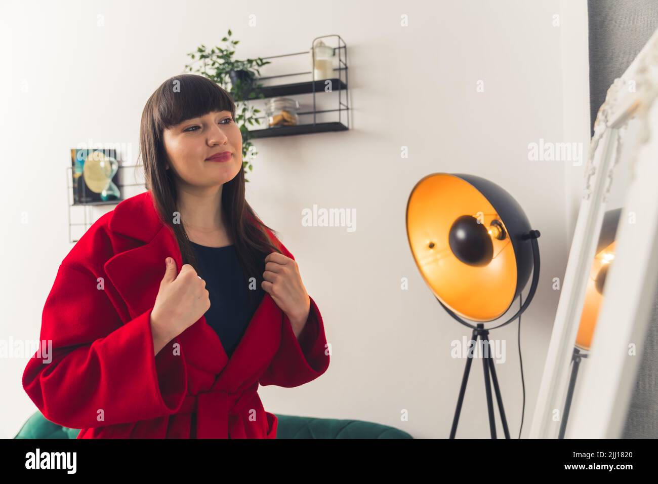 Caucasian dark-haired woman with bangs and soft makeup trying on a red coat in livingroom looking at mirror. Indoor shot. High quality photo Stock Photo