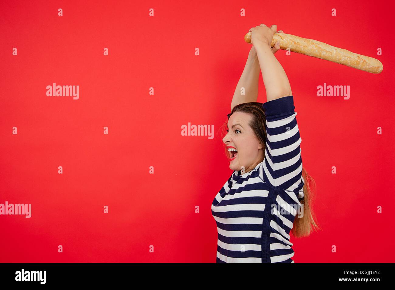 Shouting long haired plump woman in striped shirt using baguette as sword, cutting and breaking discount in red studio Stock Photo