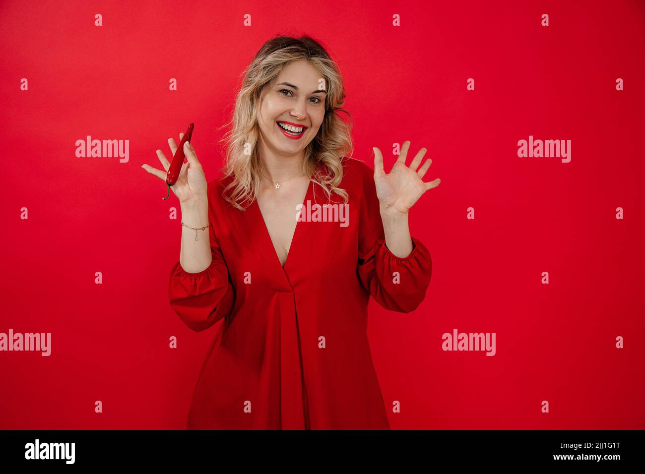 Laughing glad blond woman in red dress holding hot pepper in hand, shaking arms and gesturing, look at camera. Broadcast Stock Photo