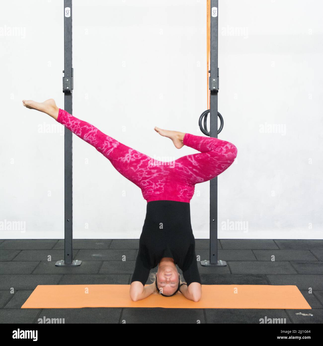 Blonde youth woman performing headstand yoga pose with feet apart but one of the legs bent. Lady wearing pink yoga leggings and black shirt in a gym. Stock Photo