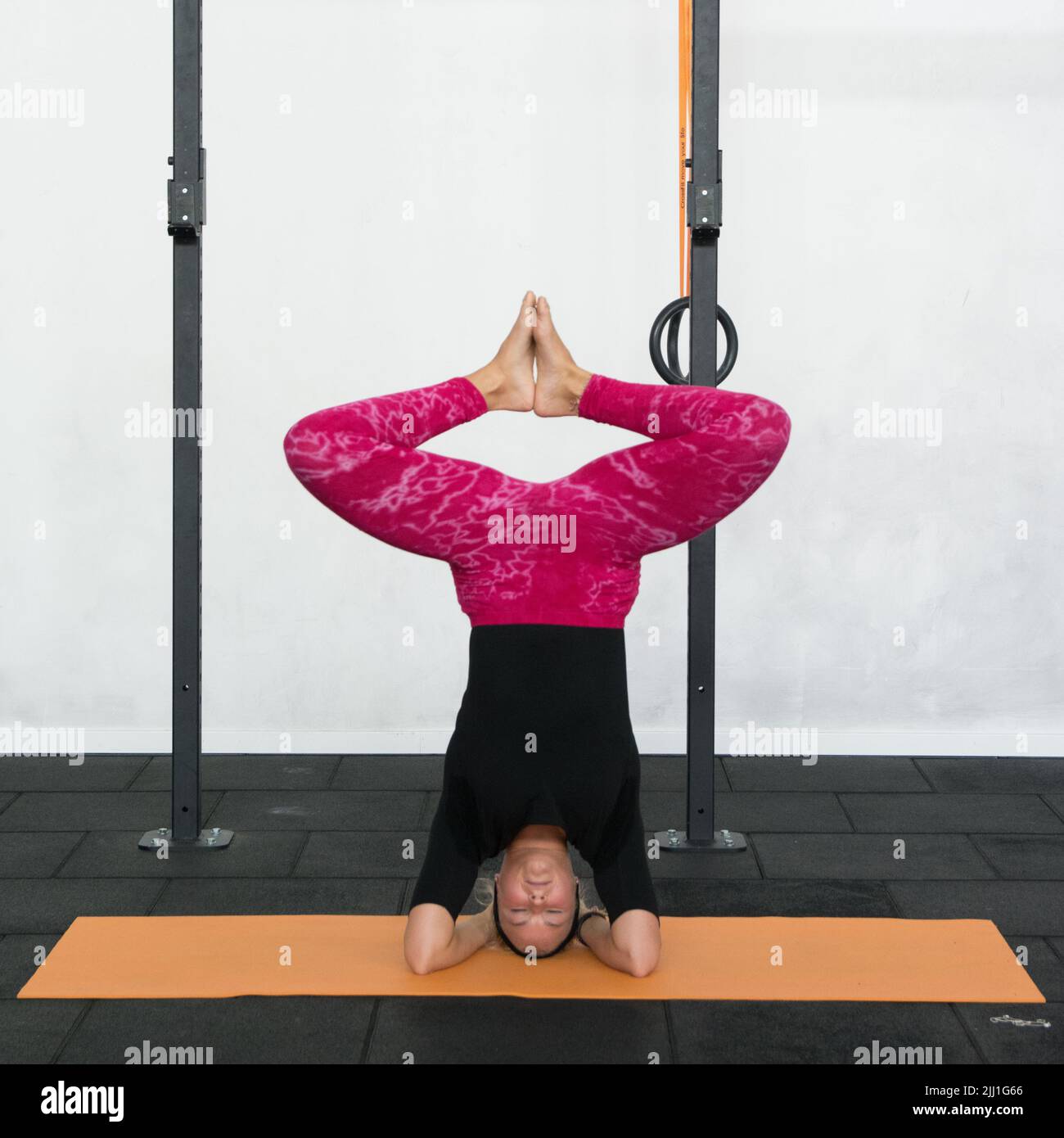 Blonde youth woman performing headstand with knees bent and foot together yoga pose wearing pink yoga leggings and black shirt in a gym. Stock Photo