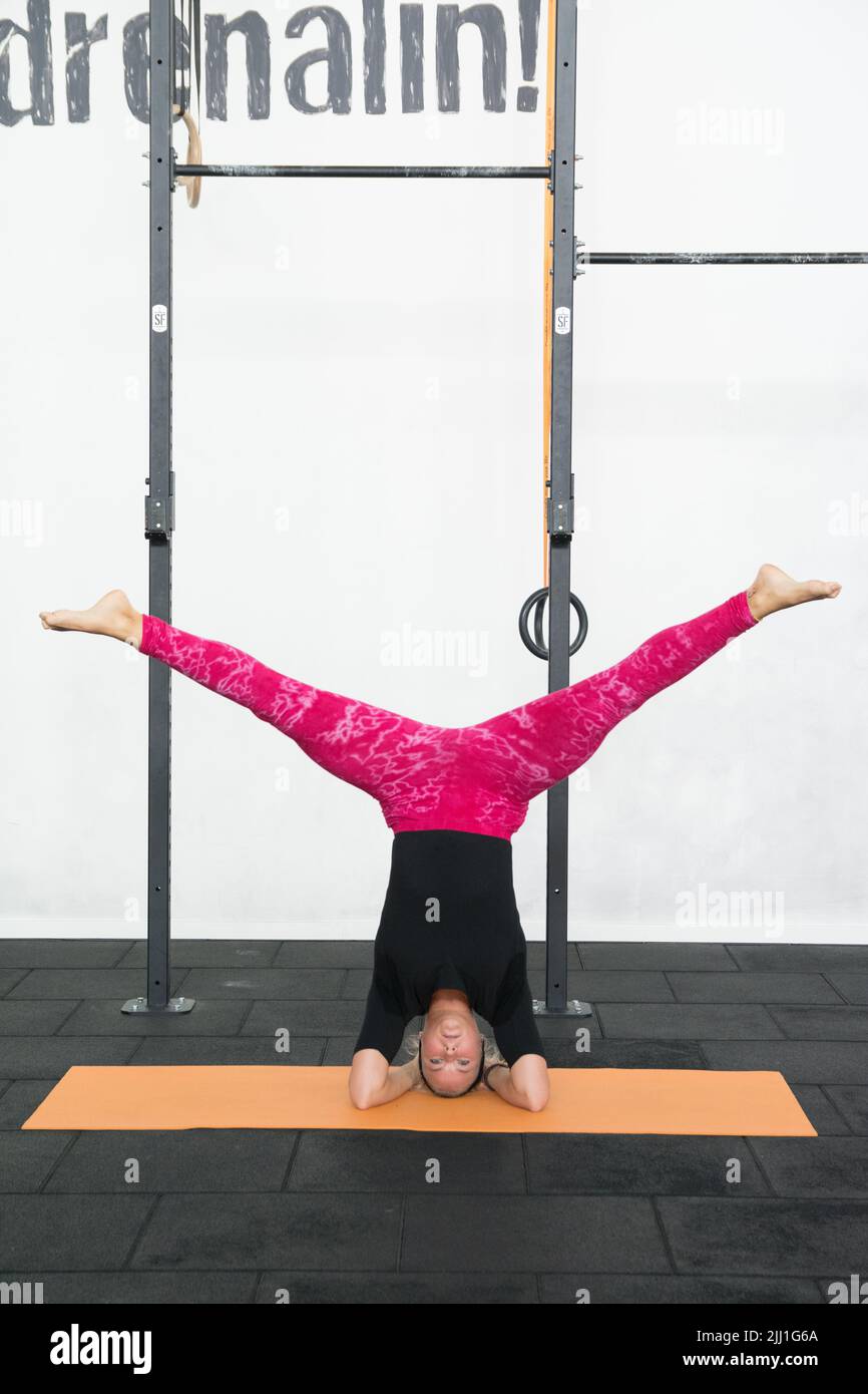 Blonde youth woman performing headstand yoga pose with feet apart wearing pink yoga leggings and black shirt in a gym. Stock Photo